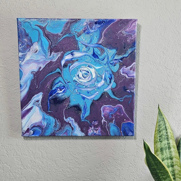 Blue Rose acrylic pour painting mounted on a neutral-toned wall, highlighting its striking blue and purple color palette with shimmering silver glitter accents.