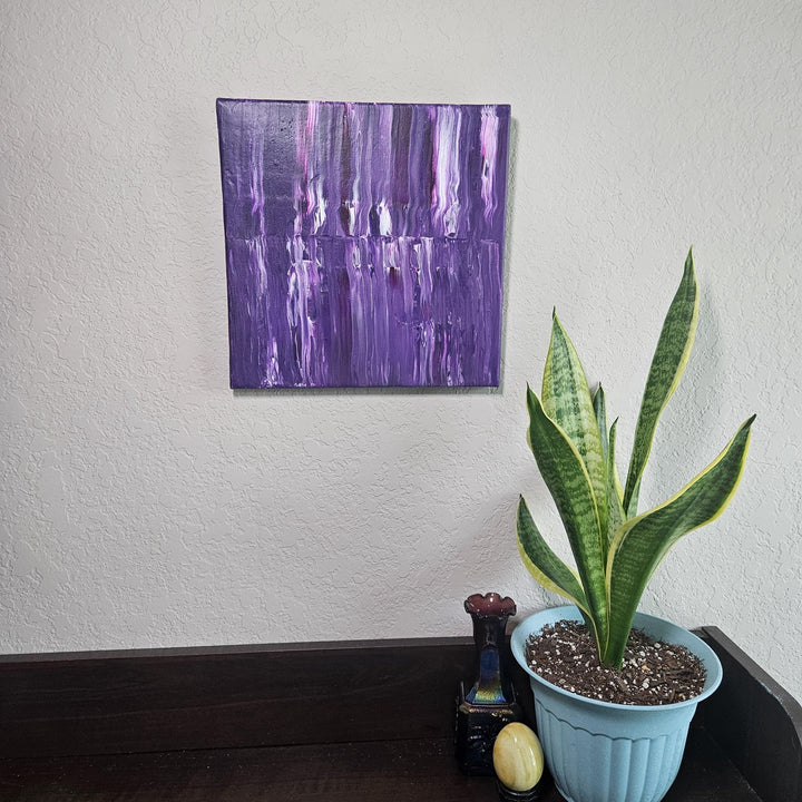 The Purple Stripe acrylic pour painting displayed on a neutral-toned wall, complemented by a plant and decorative objects, showcasing its rich violet hues and dynamic textural details.
