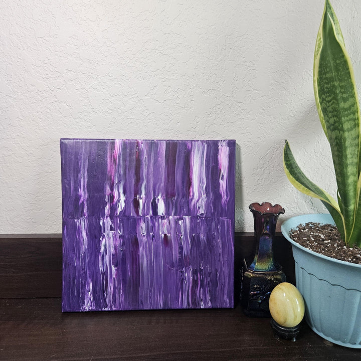 The Purple Stripe acrylic pour painting resting on a dark wooden surface, accompanied by a decorative vase and potted plant, highlighting its rich violet tones and layered texture.